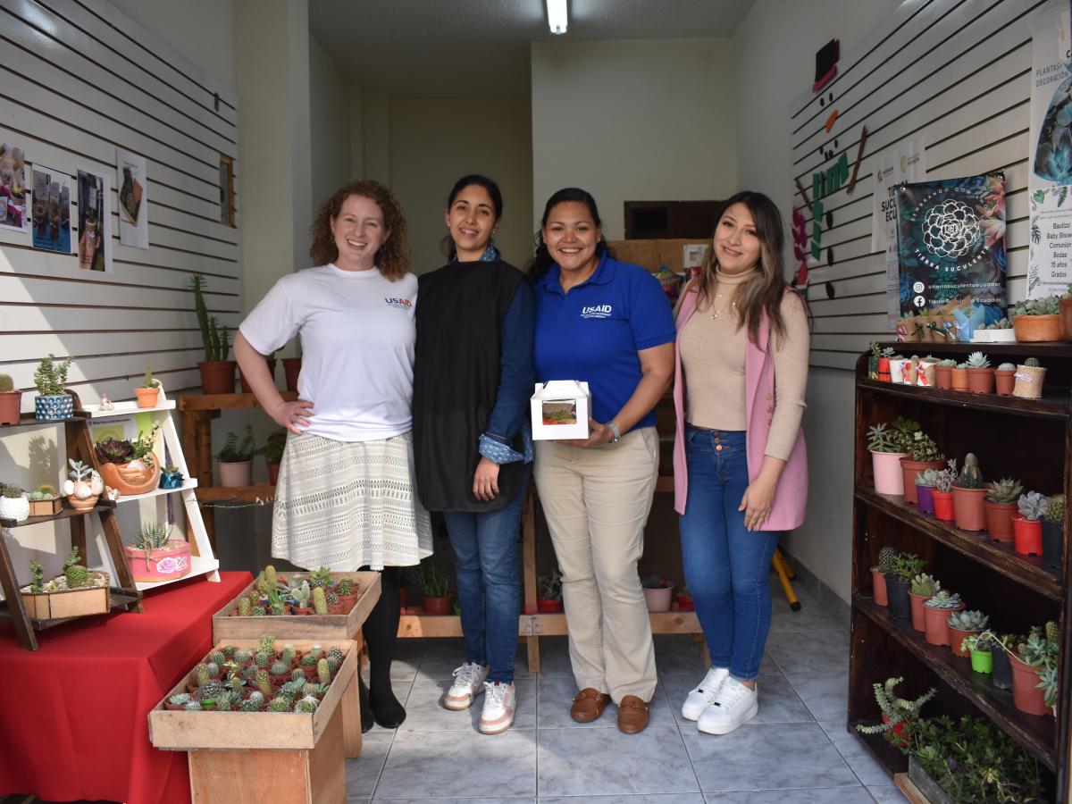 This picture shows four women standing in front of Omaira’s store. Two of the women are wearing shirts identified with the USAID logo. The other two are in informal attire, jeans and sneakers and long-sleeved shirts. 