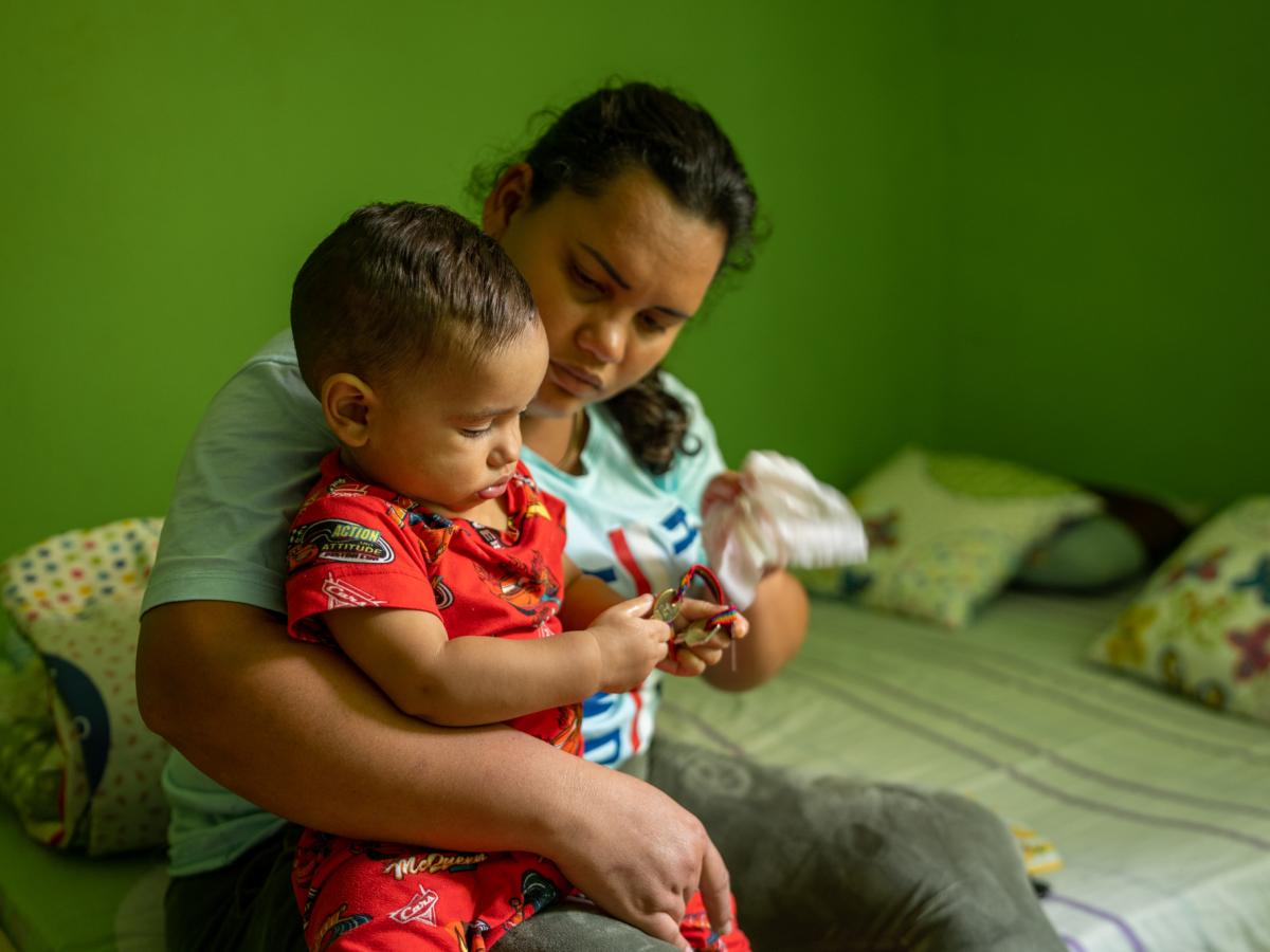 This picture shows Ariana, a female and main character in the story, hold her one-year old toddler while he plays with a bracelet. They are in a green-colored room and both are looking down. 