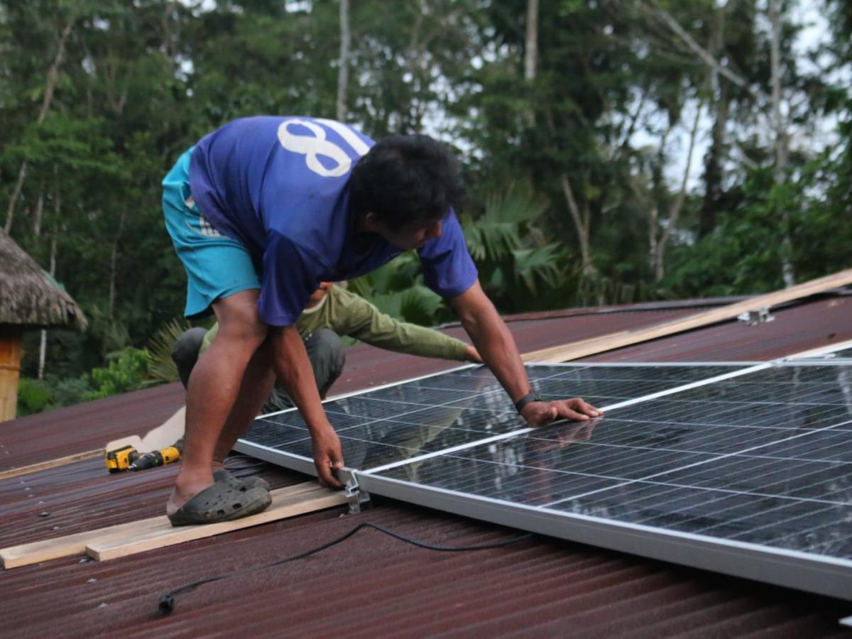 This picture shows a person, likely a young man, installing solar panels on a roof. He is in a blue t-shirt and blue shorts and is wearing comfortable shoes. There are trees on the back. 