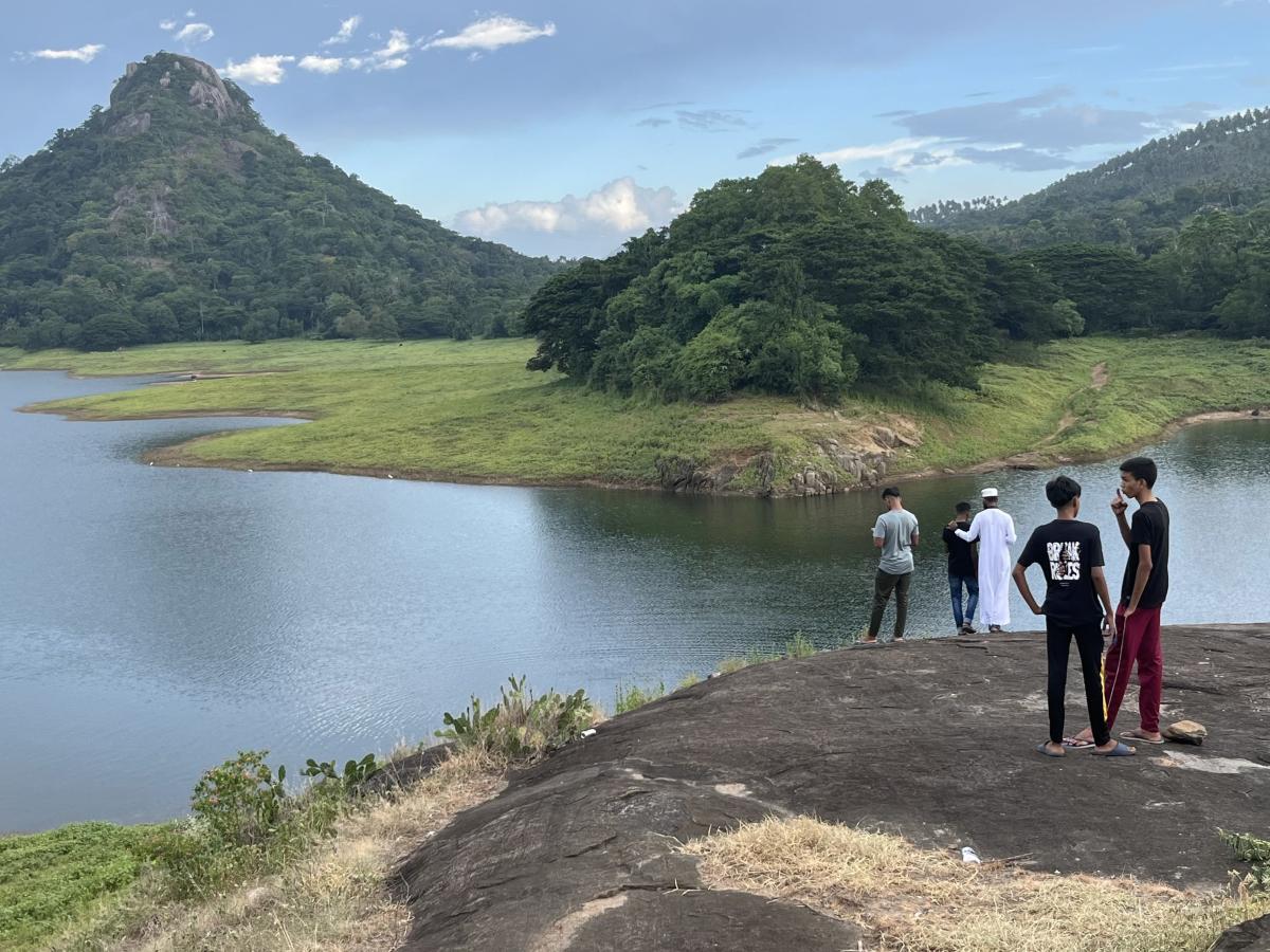 Two boys talk standing on a rock while a group of three other people look across a small lake at a mountain on the other side.