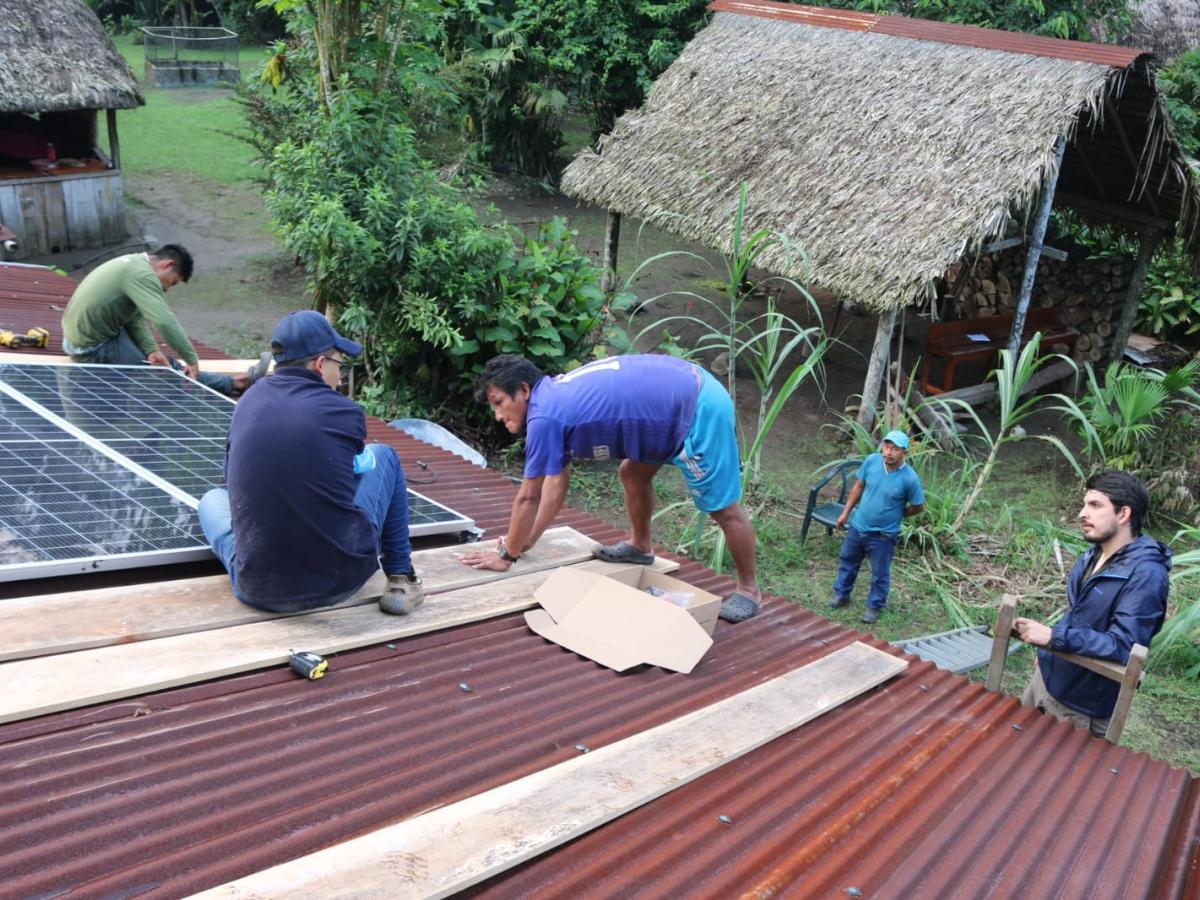 This picture shows five people, all men, installing solar panels on a roof. Two of them are sitting, standing on the roof. One of them is on a ladder, and the other standing on the ground looking up. 