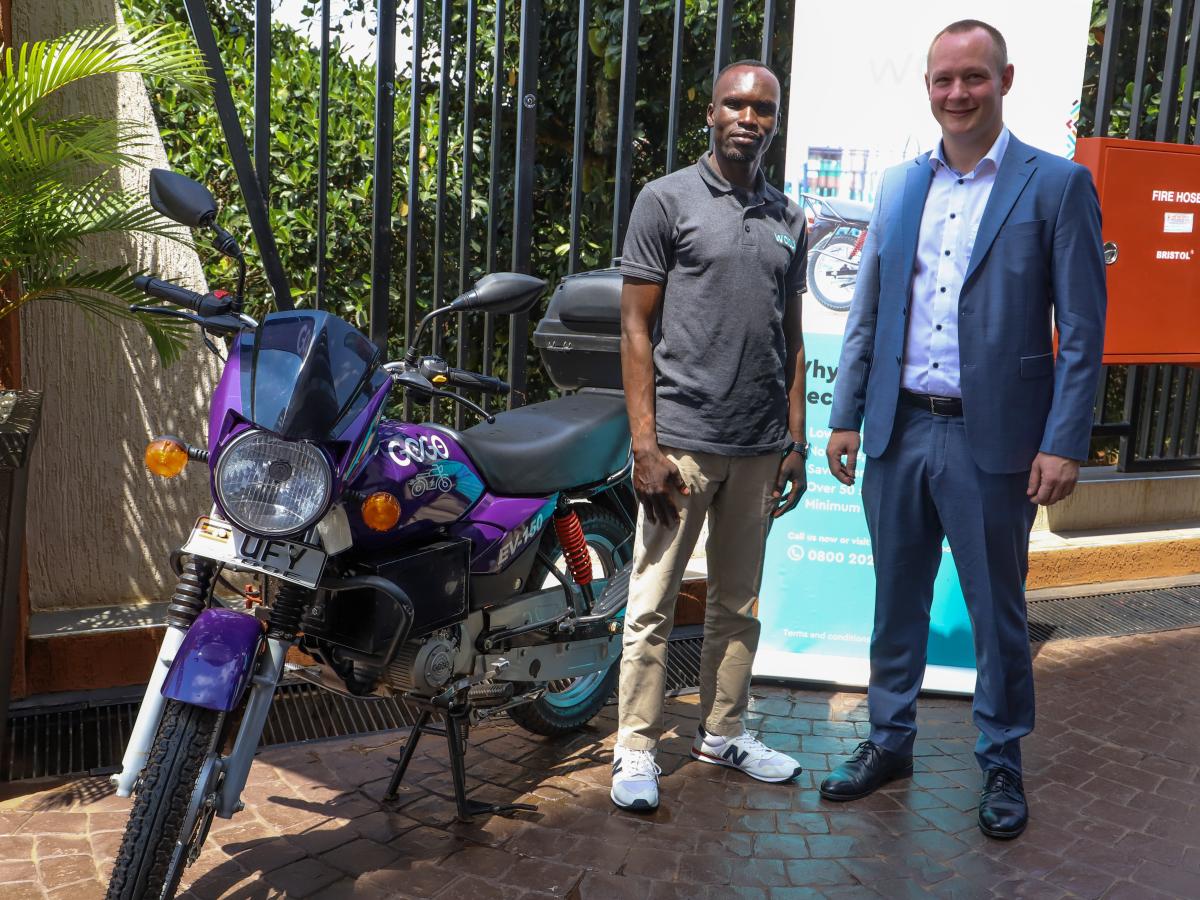 Two men stand by a purple motorbike with a banner in the background against an iron bar fence. 