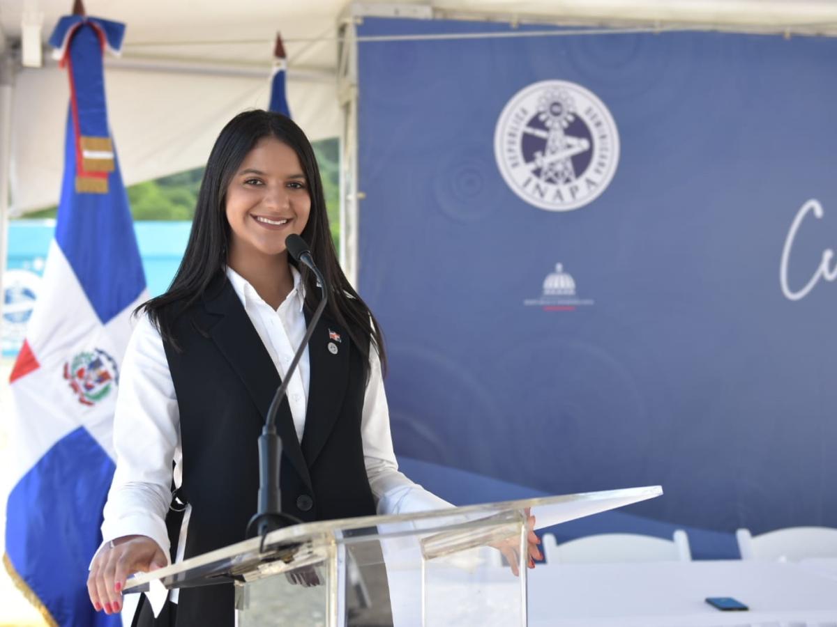 Woman speaker smiles to the camera. She is standing next to a podium.