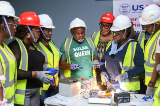 Women gather aroudn a battery and light set up, learning how solar-powered energy works