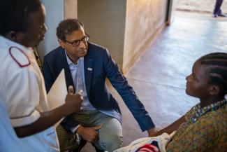 A man in a suit kneels down to speak eye to eye with a young mother holding a newborn baby on her lap.