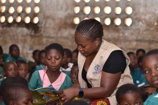 A girl in a green school uniform stands up inside her classroom to read a book while a teacher looks over her shoulder.