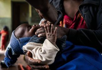 A closeup of a baby's face and hands as a an adult hand squeezes a drop of the oral polio vaccine into it's mouth.