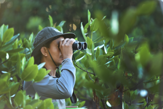 A man in a hat uses binoculars to sight waterbirds along the coast of Magalawa Island