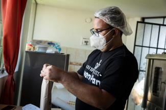 A man covering his mouth with a mask standing in a kitchen and closing a paper bag filled with bread