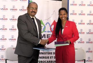 A man and a woman shake hands while exchanging documents in front of USAID and Ministry of Energy Madagascar banners. Both are smiling. 