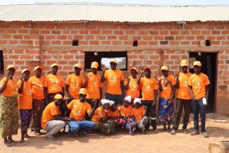 A group of 18 Zambian men and women wearing orange hats and t-shorts that read End Pandemics pose in front of a rural brick building