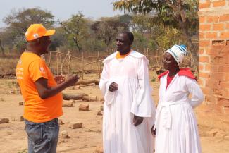 A Zambian man in a USAID t-shirt is talking to a Zambian man and woman who are dressed in the white robes of religious leaders in their church