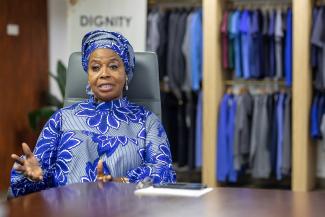 Woman dressed in blue outfit sitting behind a desk with shirts hanging behind her