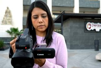 A female journalist, Jessica Ayala, stands outside a building in Mexico and holds a large black camera in her right hand as she reviews what she captured on her camera. 