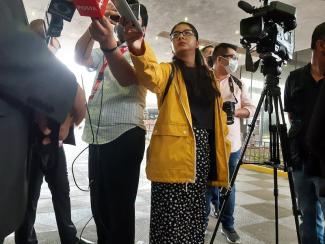 A low-angle shot of a female journalist, Ana Victoria Félix, holding up her phone to the subject as she interviews a man to the left along with a small group of reporters. 