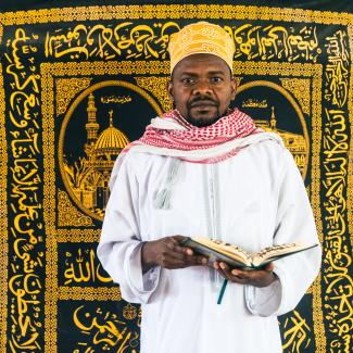  A man holding a book and standing in front of a Islamic wall art.