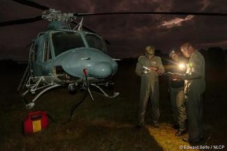 A group of 3 men stands close to a helicopter planning their journey with flashlights as they discuss the trip to transport a black rhino with a slinging technique. 