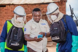 Two men wearing protective clothing and carrying backpacks and one man holding a mobile phone.