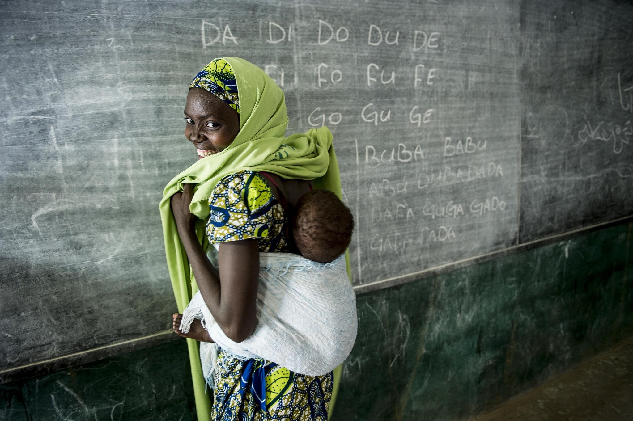 A young woman stands in front of a blackboard, infant in a sling on her back.